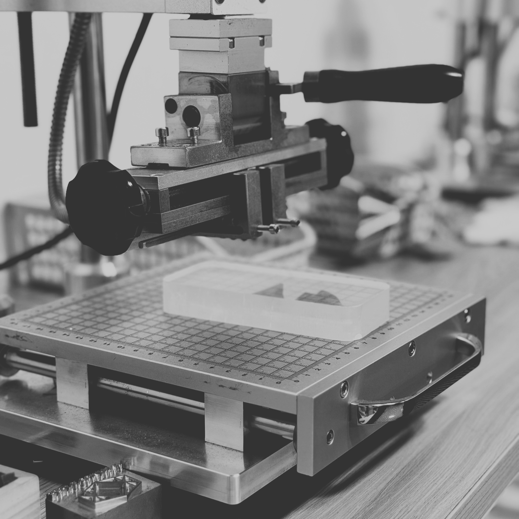 A close-up, black-and-white photograph of a desktop 3D printer or printing machine with a heated bed and a small rectangular object on the print surface.